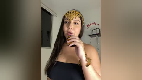 Media: Video of a woman with medium skin tone and long dark hair, wearing a gold headpiece and black strapless dress, biting her finger, in a dimly lit room.