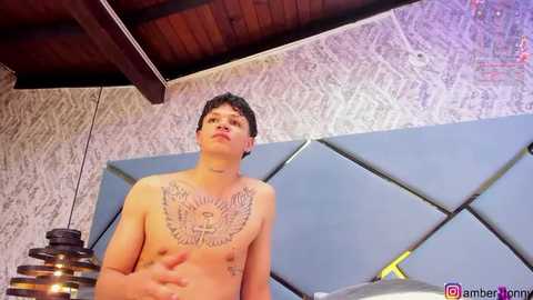 Media: Video of a shirtless, muscular, light-skinned young man with curly hair, wearing a large eagle tattoo on his chest, standing in a modern room with a geometric blue wall and wooden ceiling.