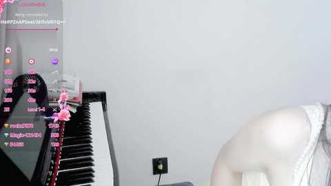 Media: Video of a woman in a white dress sitting at a piano with pink flowers, playing a melody. The background is a plain white wall with a power outlet visible.