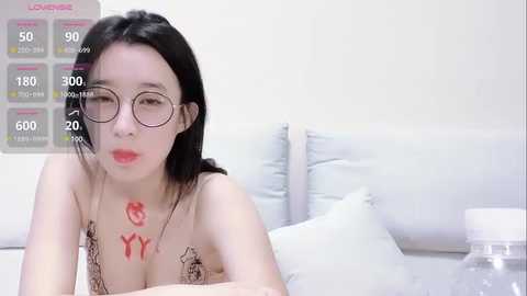 Media: Video of a young Asian woman with long black hair, wearing glasses and a nude top, lying on a white bed. She has red lipstick and a dollar sign tattoo on her chest.