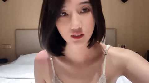 Media: Video of an Asian woman with short black hair, wearing a white lace bra, sitting on a bed with white sheets and beige headboard in a dimly lit bedroom.