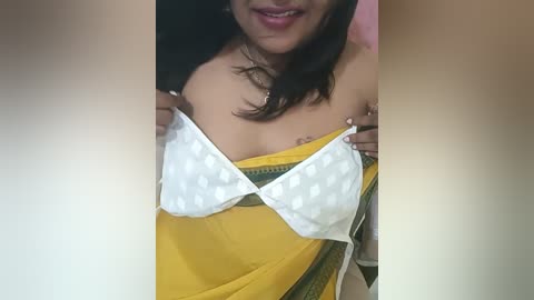 Media: Video of a South Asian woman with medium skin tone, medium breasts, and medium build, partially lifting a white polka-dot bra, revealing a yellow sari with green borders.