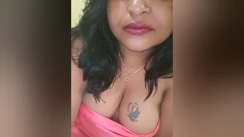 Media: A close-up video of a woman with medium brown skin, black wavy hair, and large, pink lips. She wears a pink off-shoulder top, revealing a butterfly tattoo on her left breast. The background is blurred and neutral-toned.