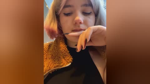 Media: Video of a young woman with light skin and platinum blonde hair, wearing a black top and brown, fuzzy jacket. She is applying eyeliner with her right hand, focusing intently.