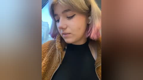 Media: Video of a young woman with light skin and shoulder-length, multi-colored hair (blonde to pink) wearing a black turtleneck and a brown fuzzy jacket. She has a contemplative expression, indoors with a blurred cityscape in the background.