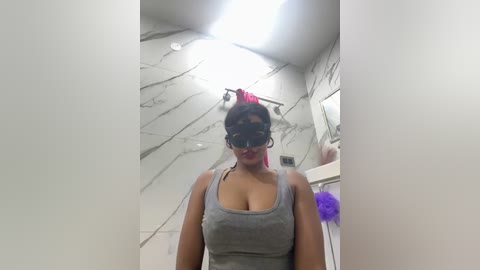 Media: Video of a woman with a medium-dark complexion, wearing a gray tank top, a black leather mask, and a red headband, standing in a bathroom with marble walls and purple cleaning supplies.