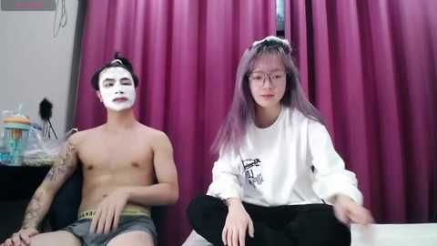 Media: Video of two Asian people, one shirtless with white face paint and a tattoo, sitting on a chair; the other wears glasses, a white sweatshirt, and black pants, sitting on a table. Background features a maroon curtain.