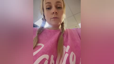 Media: Video of a young, light-skinned woman with long blonde braids, wearing a pink \"Cristiano\" t-shirt, standing indoors with a blurred, pink gradient background.