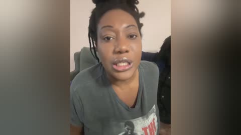 Media: Video of a young Black woman with medium brown skin, wearing a grey T-shirt, sitting indoors. She has short, curly hair styled in a bun and a neutral expression.