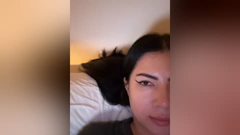 Media: Video of a woman with long black hair, fair skin, and dark eyeliner, lying on a white bed in a dimly lit room, partially obscured by a blurred, beige wall.