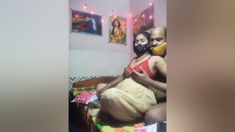 Media: A video of a couple in a small, dimly lit room. The woman, wearing a yellow sari and black mask, sits on a bed with colorful sheets, while a bald man, wearing a red shirt, embraces her from behind. The room features religious art and pink fairy lights.