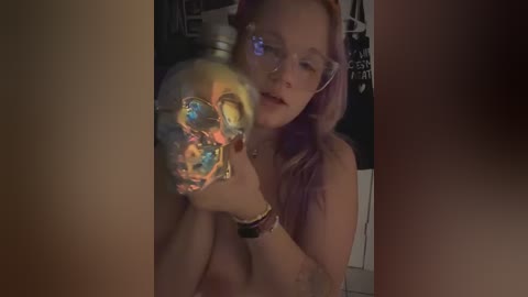 Media: Video of a topless woman with light skin and pink hair, holding a reflective, colorful skull-shaped light. Background shows dimly lit room with white shelves.