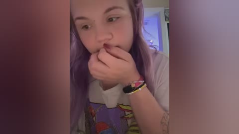 Media: Video of a young girl with light skin, long lavender hair, and a white T-shirt featuring a cartoon character. She is holding her fingers to her lips, in a blurred, intimate setting.