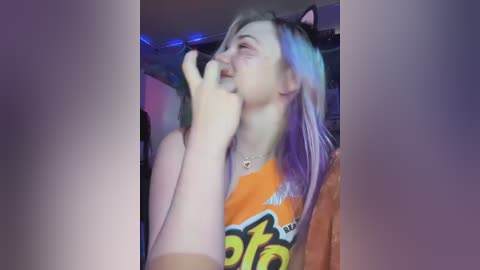 Media: Video of a young woman with long, platinum blonde hair, wearing a colorful orange T-shirt, smiling with her fingers near her mouth, in a dimly lit room with blue LED strip lights.