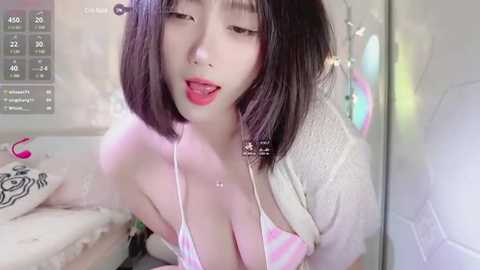 Media: A young East Asian woman with short black hair, wearing a low-cut pink and white striped bikini top, and a white cardigan, is seen indoors with a playful, slightly provocative expression.