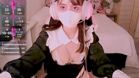 Media: A video of an Asian woman wearing a revealing black and white French maid outfit with a white mask and headphones, sitting on a bed with a pink background.