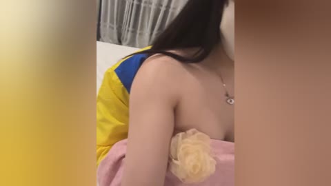 Media: Video of a young woman with long black hair, wearing a yellow and blue sports jacket, partially covering her bare shoulders. She is seated, holding a large yellow bath sponge. Background features a white curtain and a pink towel.