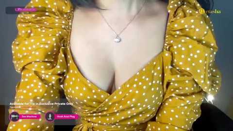 Media: Video of a woman with fair skin wearing a low-cut mustard-yellow dress with white polka dots, showcasing cleavage, and a delicate necklace. Background is blurred.