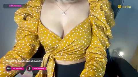 Media: Video of a woman with fair skin and medium-sized breasts wearing a mustard-yellow polka-dotted top tied at the waist, revealing cleavage. She is seated, indoors, with a background showing a chair and a lamp.