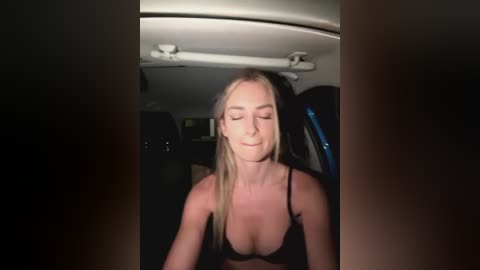 Media: Video of a blonde woman with fair skin, wearing a black bra, smiling, sitting in a car with dim interior lighting.