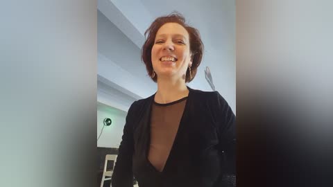 Media: Video of a smiling woman with short brown hair, wearing a black velvet jacket over a sheer black top, standing in a modern, brightly lit room with white walls and a green light fixture.