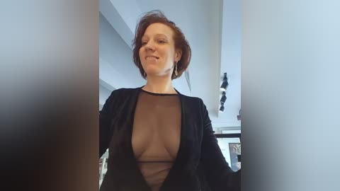 Media: Video of a smiling, middle-aged woman with short, auburn hair, wearing a sheer black top revealing her cleavage, in a modern, brightly lit room with white walls and minimalistic decor.