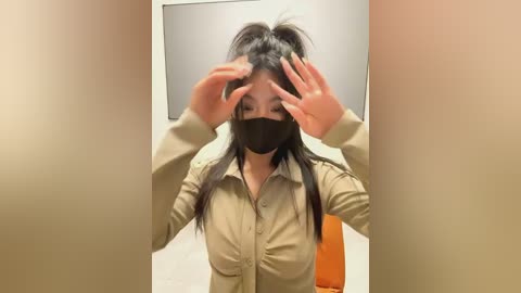 Media: Video of a young Asian woman with long black hair, wearing a beige button-up shirt, black face mask, and orange chair, adjusting her hair in a small, beige-walled room.