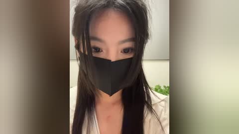 Media: A video of an East Asian woman with long black hair, wearing a black face mask and white shirt, against a blurred beige background.