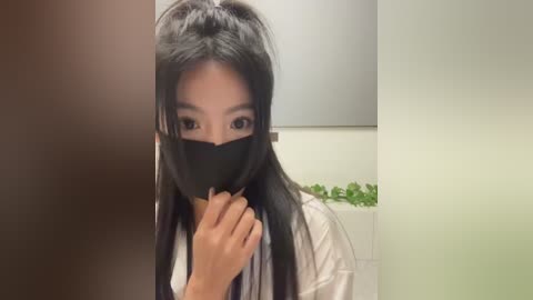 Media: Video of an Asian woman with long black hair and fair skin, wearing a black face mask and white lab coat, holding a syringe, indoors with blurred background.