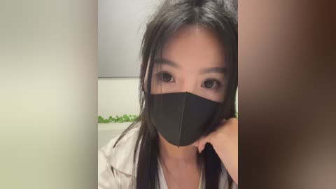 Media: Video of an East Asian woman with long black hair, wearing a black face mask and light jacket, taken indoors against a blurred background.