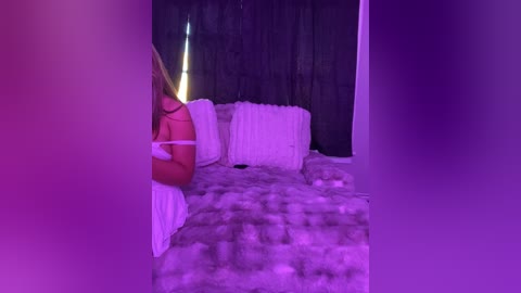 Media: Video of a dimly lit bedroom with purple lighting. A blonde woman in lingerie is partially visible, sitting on a white fluffy bed with white pillows. Black curtains cover the background.