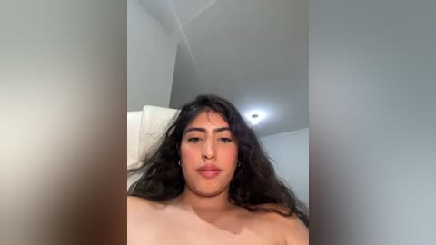 Media: A video of a topless woman with long, wavy black hair, light brown skin, and a full nose, lying on a bed. The background features a white ceiling with a light fixture and a white wall.