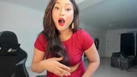 Media: Video of a South Asian woman with long, wavy brown hair and fair skin, wearing a red top, standing in a modern, brightly lit living room. She has a surprised expression with open mouth and wide eyes.