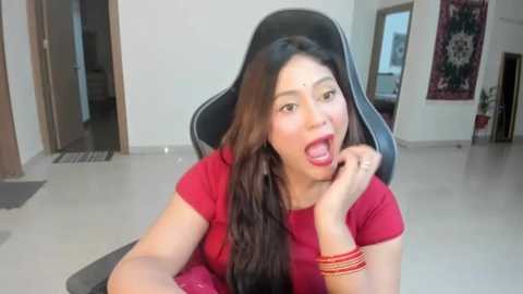 Media: Video of an Asian woman with long black hair, wearing a red top and multiple bracelets, sitting in a gaming chair, open-mouthed, in a modern, brightly lit room with white walls and a door in the background.