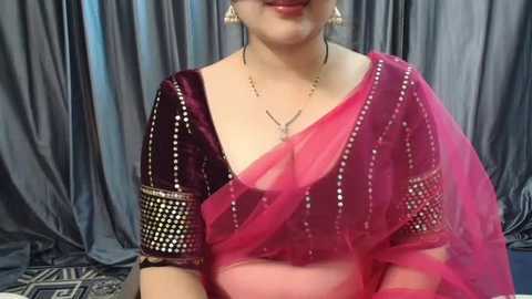 Media: Video of a woman in a deep pink sari with intricate gold embroidery on the sleeves, paired with a sheer pink dupatta. She wears gold earrings and a delicate necklace. Gray drapes form the background.