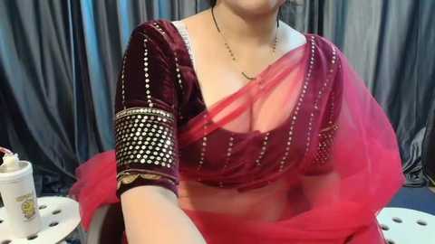 Media: Video of a South Asian woman in a maroon velvet blouse and red saree, adorned with gold sequins, seated on a white chair. The background features dark grey curtains.