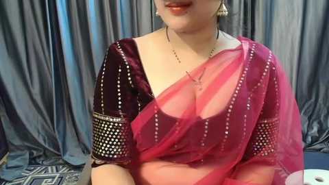 Media: Video of a South Asian woman in a deep red sari with gold sequin embroidery, adorned with a gold necklace and dangling earrings. She sits against gray drapes in a formal setting.