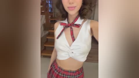 Media: Video of a woman in a tight, white sleeveless crop top with a red bow, revealing midriff, and matching red plaid skirt. Background shows wooden stairs and a beige wall.