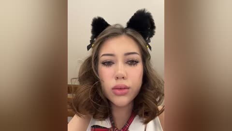 Media: Video of a young woman with fair skin, wearing black cat ears and a white shirt, with a neutral expression and wavy brown hair.