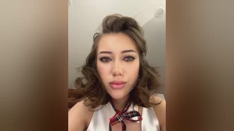 Media: Video of a young woman with wavy brown hair, wearing a white sleeveless top and a red and black plaid tie. She has light skin, full lips, and is indoors against a plain, beige background.