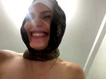 Media: A video shows a topless woman with light skin, wearing a black lace hood with a large, shiny, reflective surface. She has a bright smile and closed eyes. The background is a plain, light-colored wall.