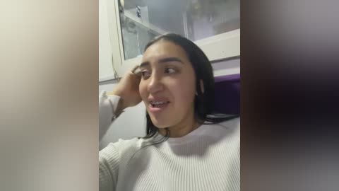 Media: Video of a young woman with long black hair, fair skin, and braces, wearing a white ribbed sweater, sitting in a purple chair, looking out a window, with a blurred background.