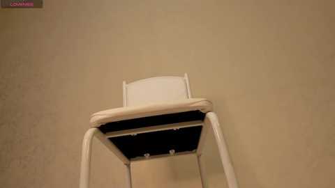 Media: Video of a white plastic chair with a black seat and backrest against a beige textured wall. The chair appears empty, and the image is taken from a low angle, emphasizing the chair's height.