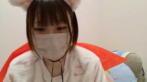 Media: Video of an Asian woman with straight black hair and bangs, wearing a white surgical mask, a fluffy white hat with ears, and a white robe. She sits on a red and blue chair against a plain white wall, looking down.