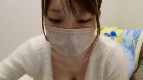 Media: Video of an East Asian woman with light skin and dark hair, wearing a white mask and fluffy robe, lying on a bed with colorful pillows.