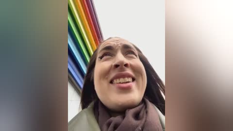 Media: Video of a smiling woman with long dark hair and fair skin, wearing a brown scarf and jacket, against a backdrop of a rainbow-striped mural.