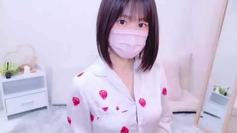 Media: Video of an East Asian woman with shoulder-length dark hair, wearing a white button-up shirt with red strawberry patterns, a pink face mask, and a white surgical mask, standing in a minimalist white room with a white chair and a small white table.