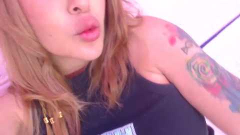 Media: Video of a light-skinned woman with long, wavy, blonde hair, puckering her lips. She has a black sleeveless top and a colorful tattoo on her right arm. The background is blurry, likely indoors.