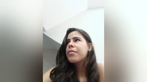 Media: A video of a young woman with long, wavy black hair, light skin, and a neutral expression, standing indoors with a white ceiling and beige walls.