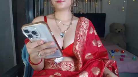 Media: Video of a woman in a red sari with gold embroidery, wearing a black necklace and earrings, holding a smartphone, in a cozy room with a teddy bear and colorful makeup brushes.